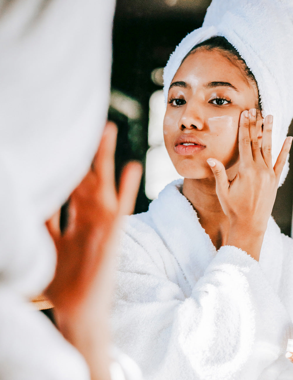 Woman putting natural anti-aging cream on her face in mirror wearing a bathrobe and towel on her head