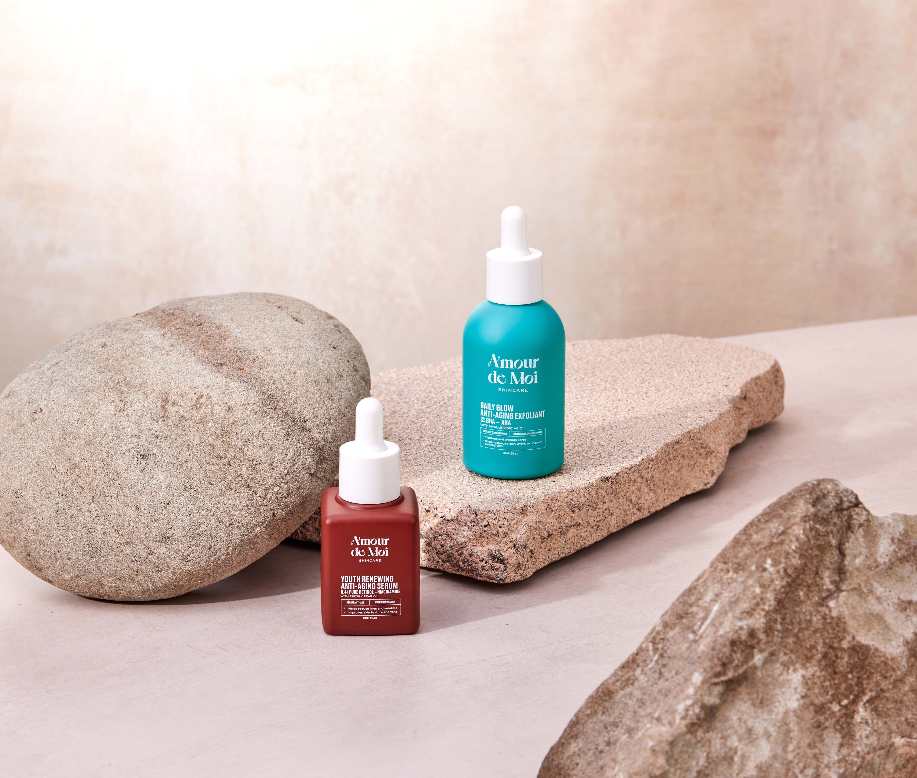 a bottle of the youth renewing anti-aging serum and the BHA AHA daily glow anti-aging exfoliant sitting on rocks