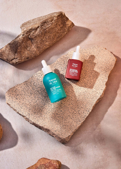 a bottle of the youth renewing anti-aging serum and the BHA AHA daily glow anti-aging exfoliant laying on rocks