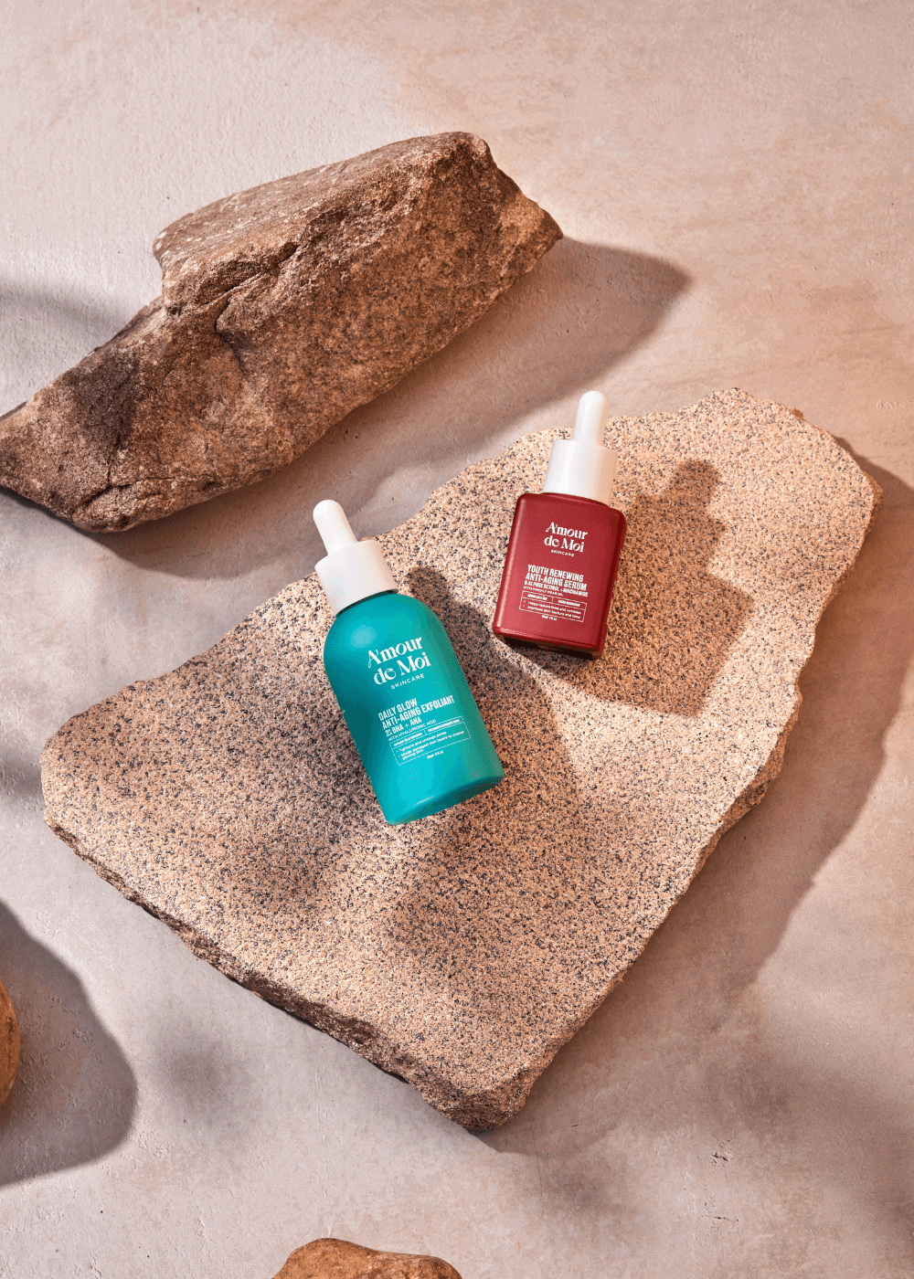 a bottle of the youth renewing anti-aging serum and the BHA AHA daily glow anti-aging exfoliant laying on rocks